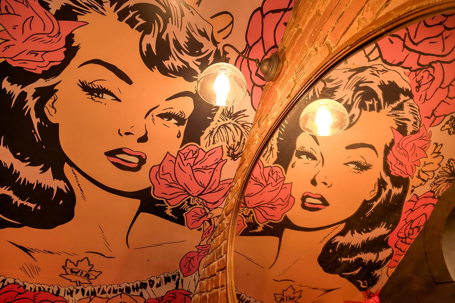 Pop art style mural of a woman with dark hair, red lips, and a tear, decorated with pink roses, reflected in an oval mirror with a wall light above.