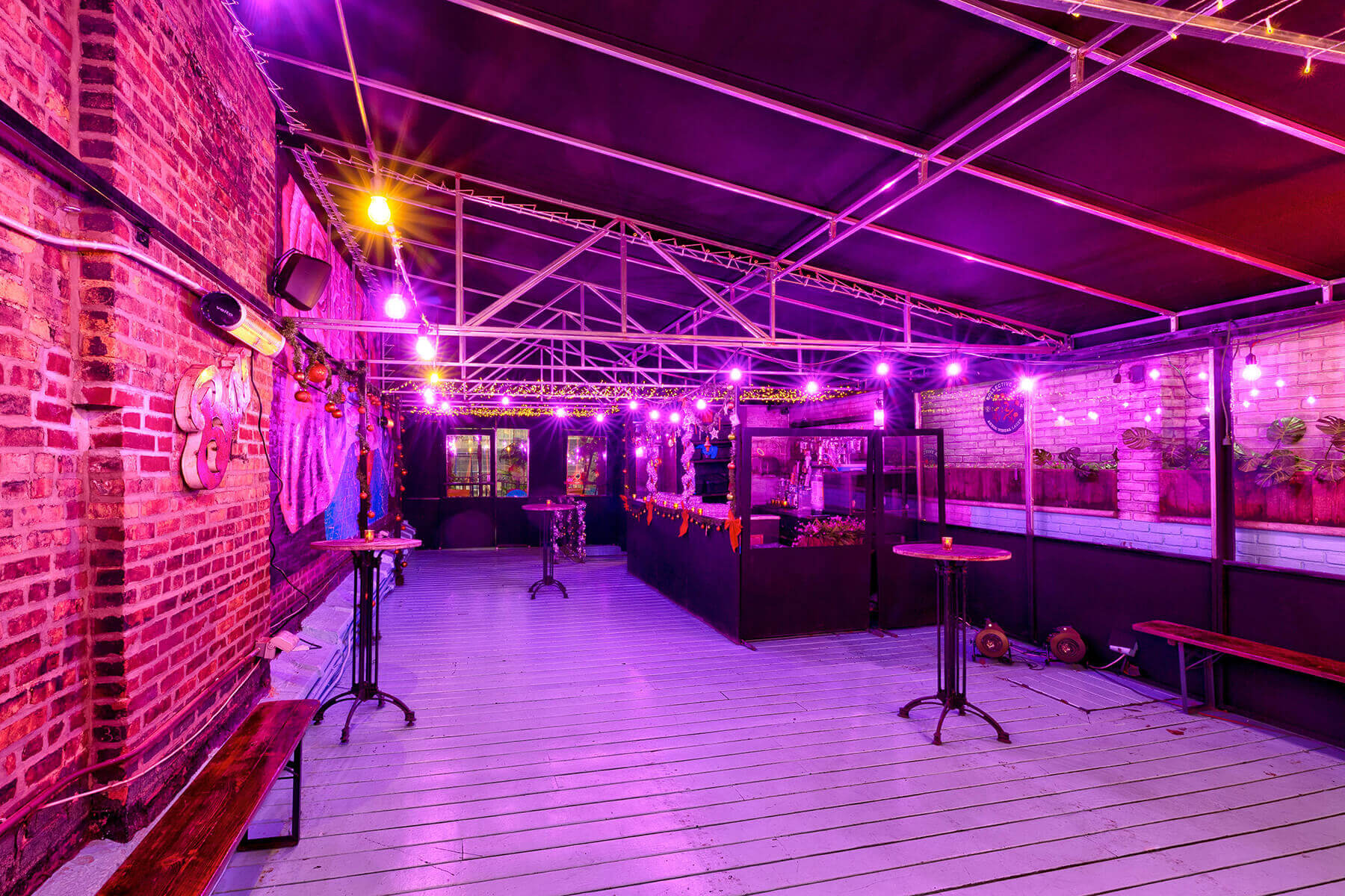 Indoor bar area with brick walls, purple string lights, high tables, and benches under a black ceiling with metal beams.