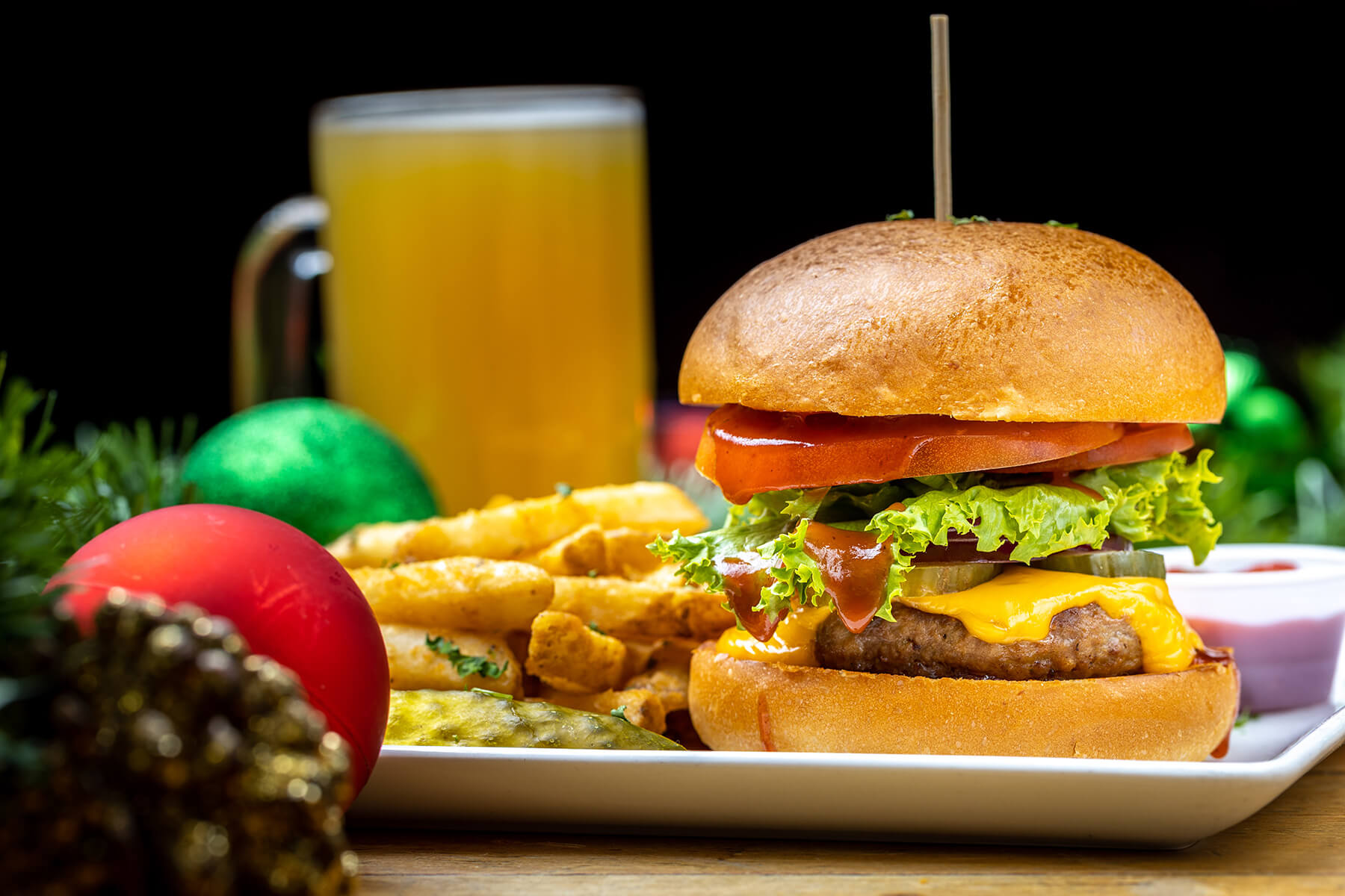 Cheeseburger with lettuce, tomato, pickles, and ketchup on a bun served with crinkle-cut fries, a pickle, dipping sauce, and a glass of beer.