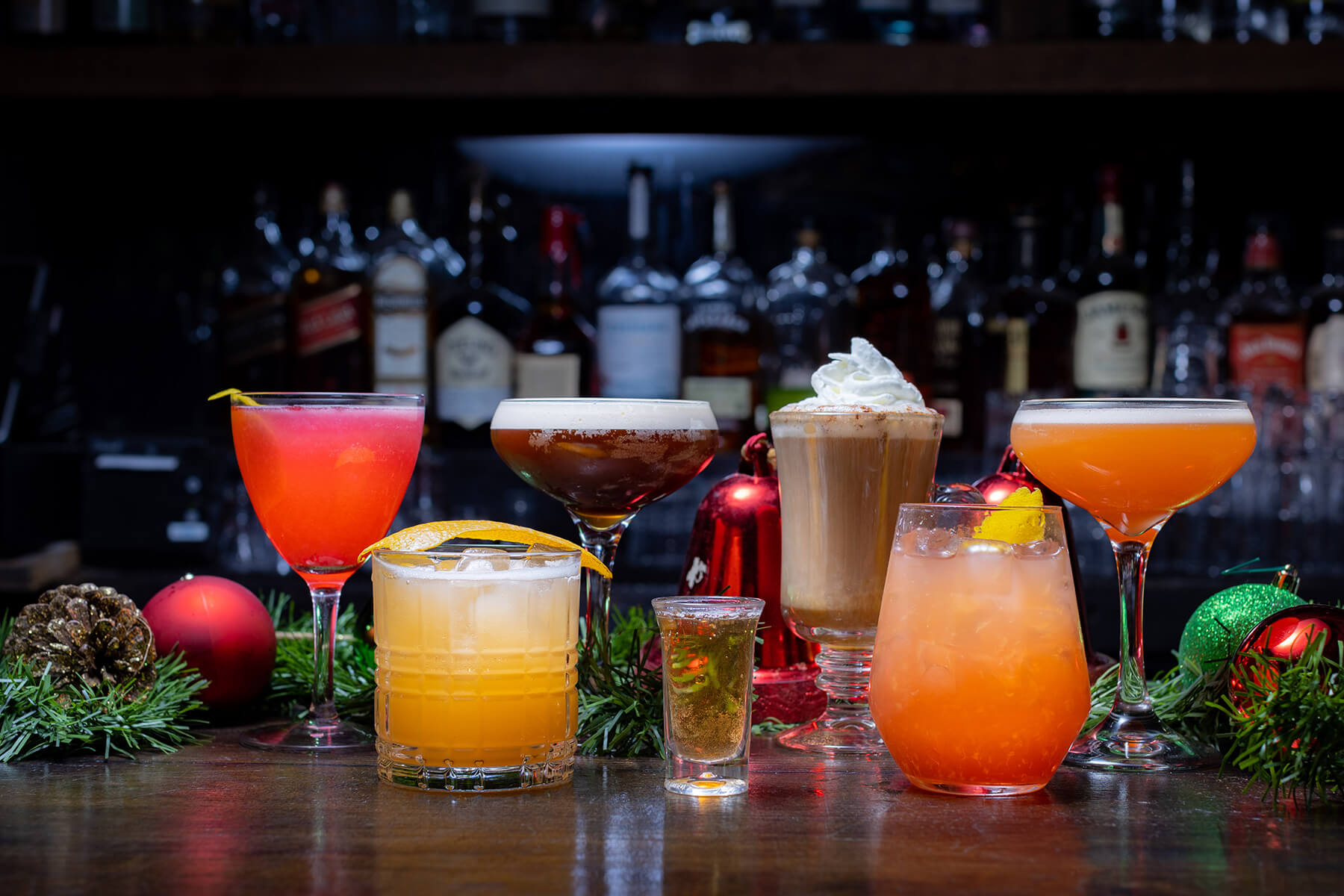 Six colorful cocktails arranged on a bar counter with festive holiday decorations and liquor bottles in the background.