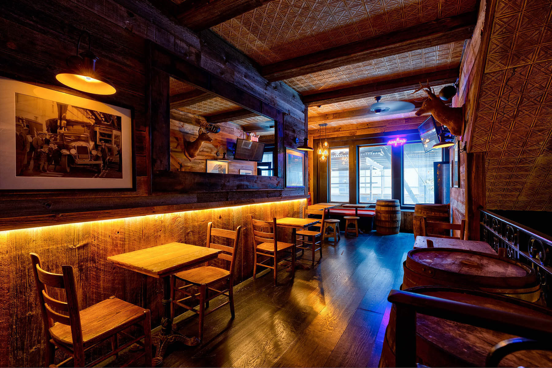 Cozy rustic pub interior with wooden tables, chairs, barrel decor, mounted deer heads, a large mirror, and warm yellow lighting.