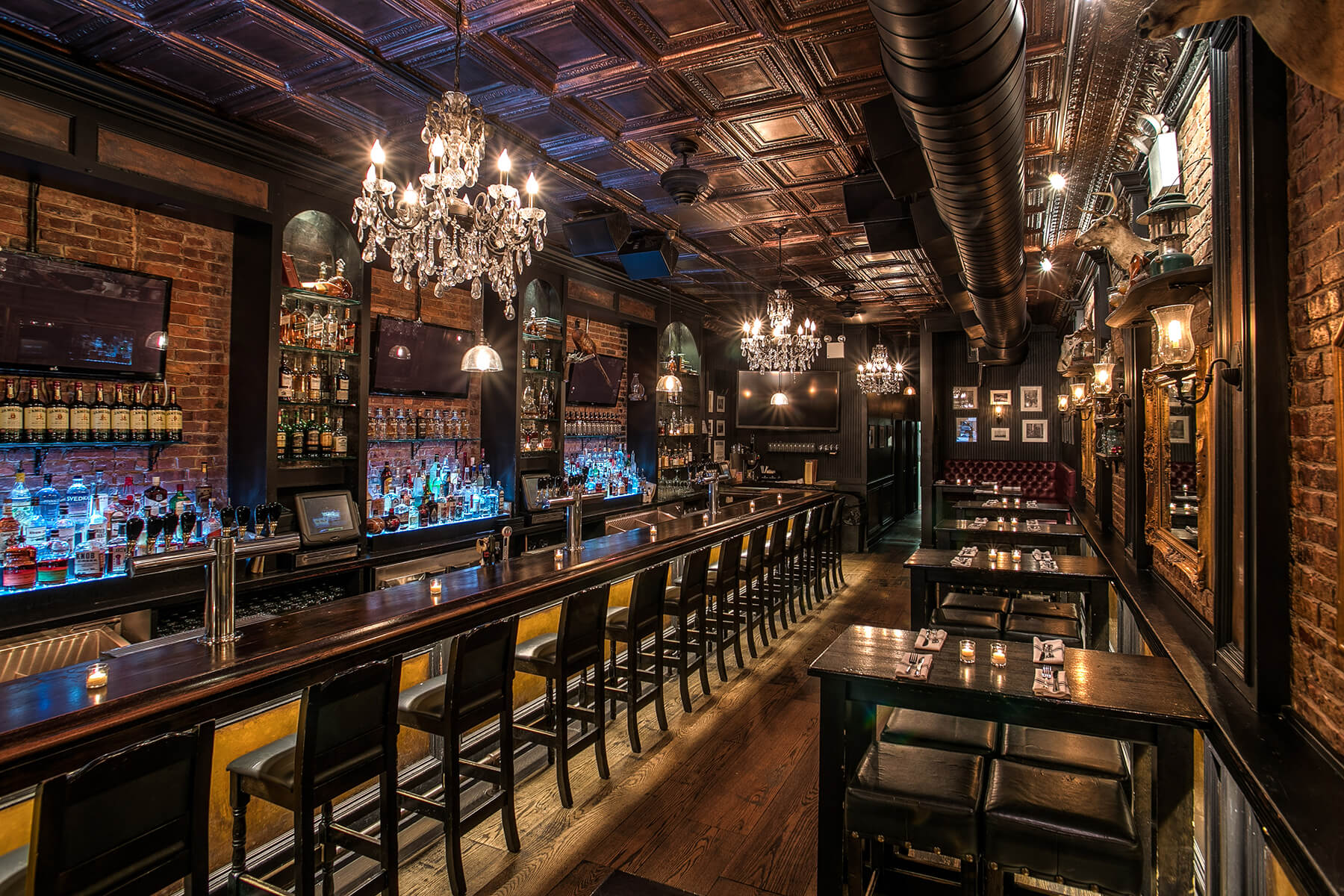 Elegant bar interior with a long wooden counter, chandelier lighting, exposed brick walls, and rows of bottles on shelves behind the bar.